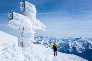 A skier walks past a snow and ice covered cross atop a mountain in front of a mountain range and blue skies