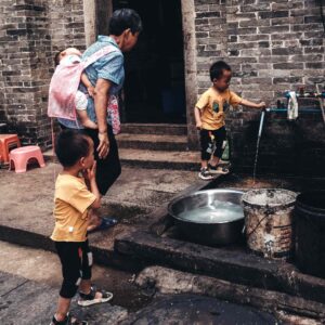 a parents supervises two young boys fetching water from an outdoor tap